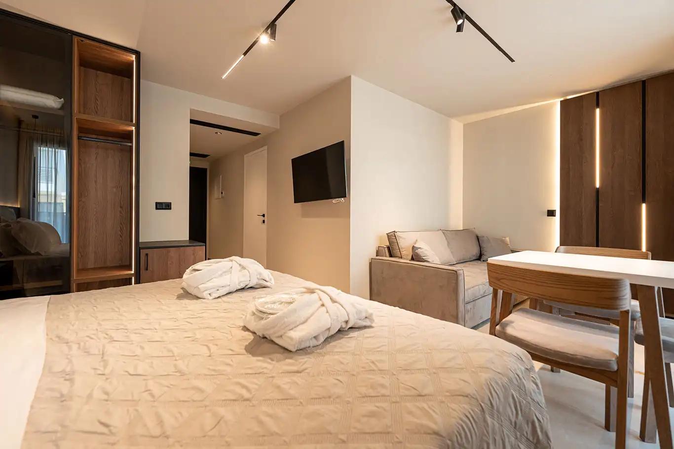 Double bed with bathrobes overlooking a sofa, dining table, television and illuminated wood panels