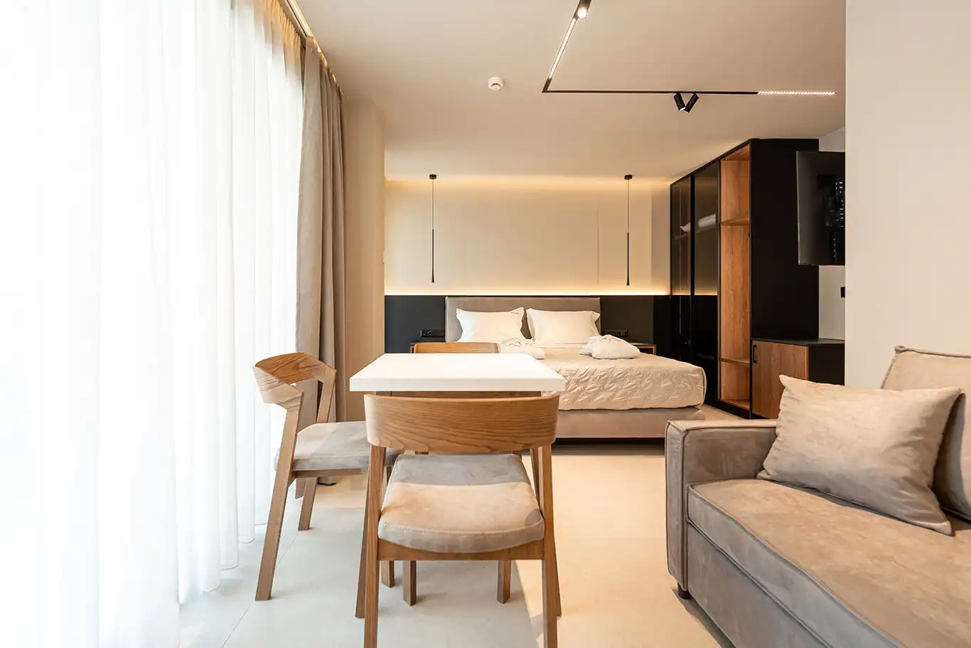 Open-plan interior with double bed, white dining table, wooden chairs, sofa and built-in wardrobe