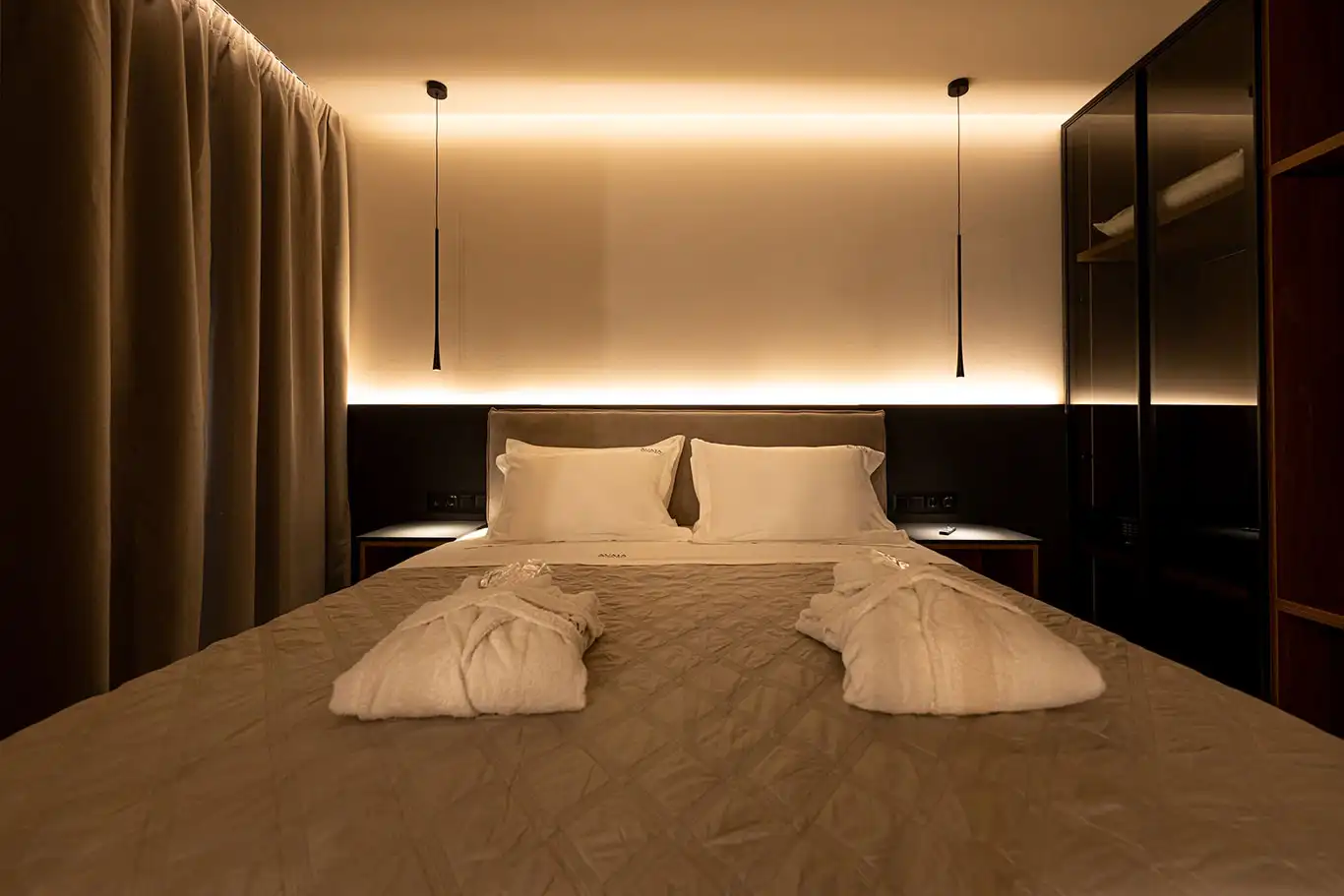 Front view of a double bed with white bedding, folded bathrobes, pendant lights and dark glass wardrobe