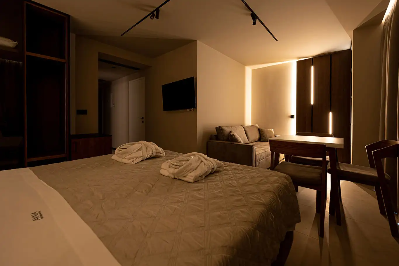 Wide interior of Adjacent Deluxe Luxury Room 104 with double bed, bathrobes, sofa, dining table and television