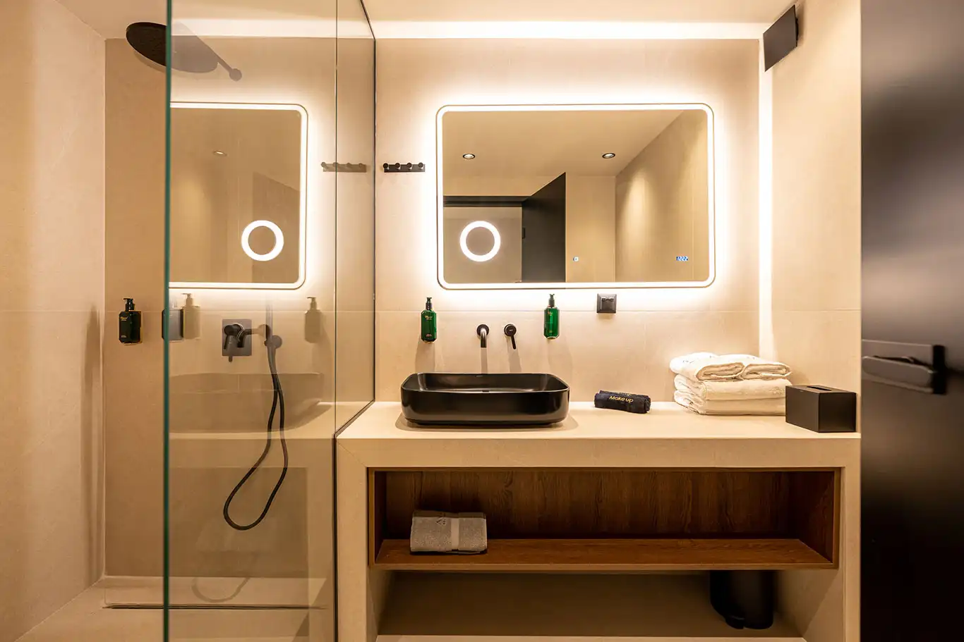 Spacious bathroom vanity with black basin, illuminated mirror, folded towels and adjacent glass shower
