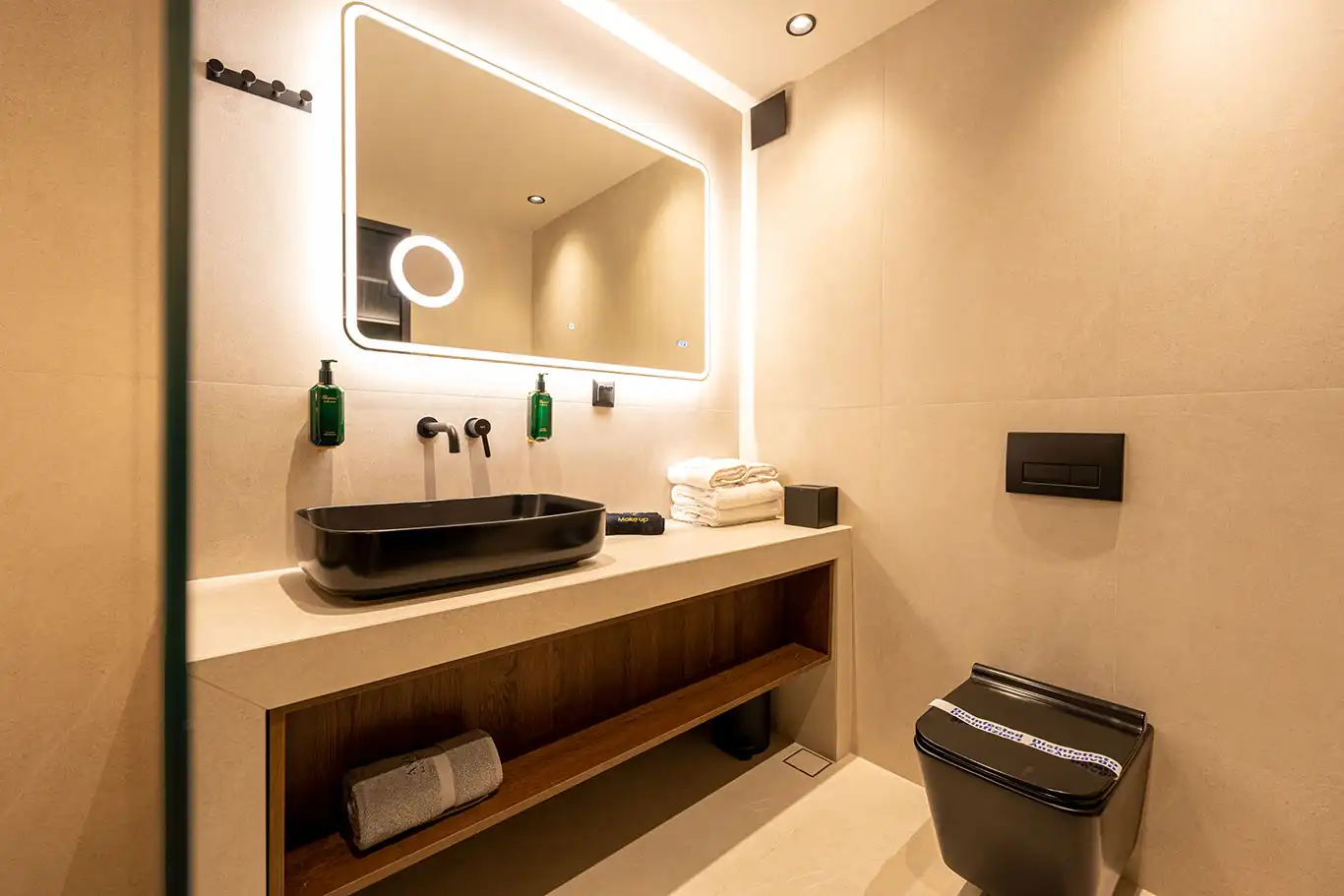 Modern bathroom with black countertop basin, illuminated mirror, folded towels and black wall-mounted toilet