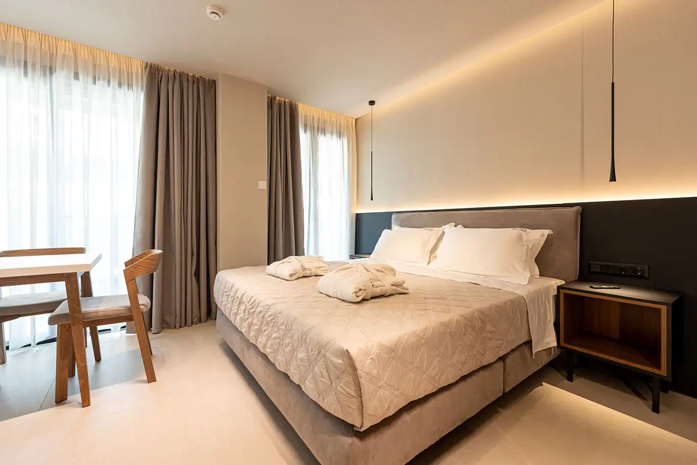 Double bed with white bedding, bathrobes, dining table, wooden chairs and bright windows