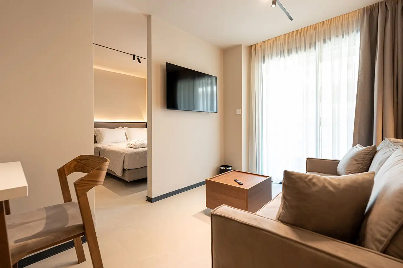 Bright living area with beige sofa, wooden coffee table, wall-mounted television and doorway leading to the bedroom in Adjacent Deluxe Luxury Room 105