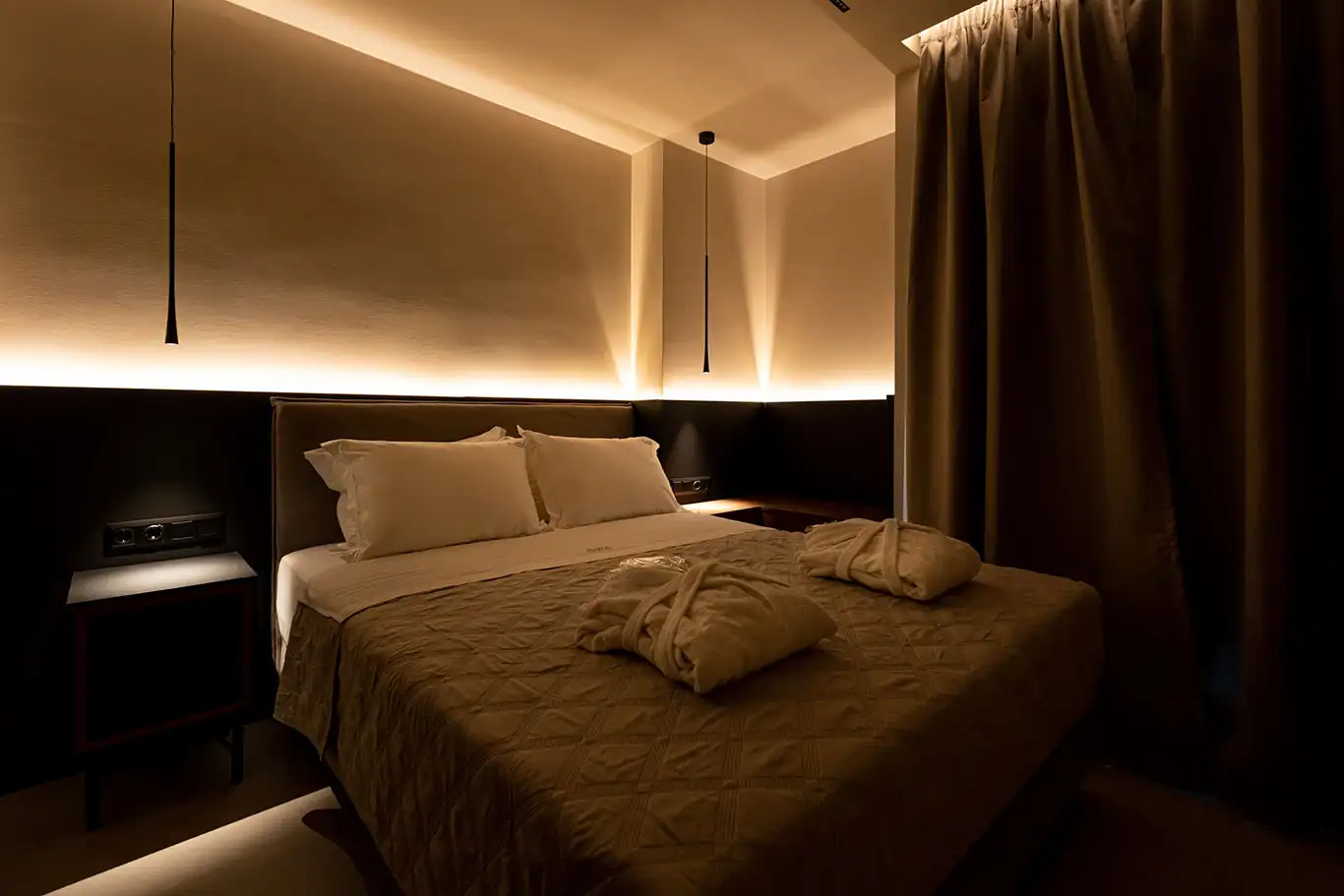 Double bed with white pillows, folded bathrobes, bedside tables, pendant lights and dark curtains
