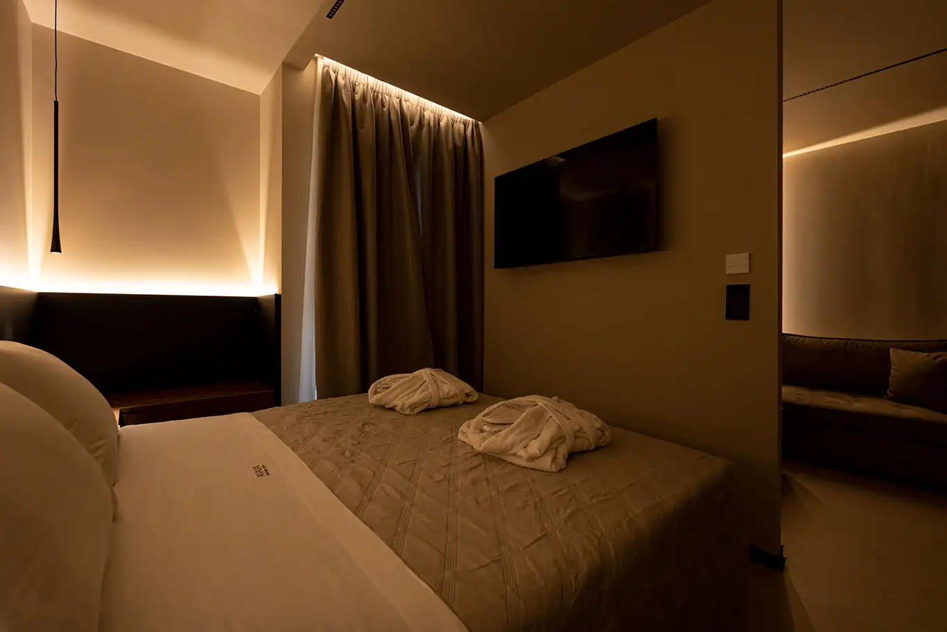 Double bed with folded bathrobes, wall-mounted television and warm ambient lighting in Adjacent Deluxe Luxury Room 105