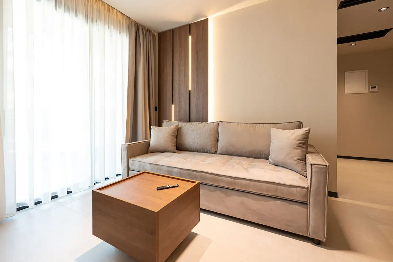 Spacious beige sofa with decorative cushions beside a wooden coffee table and large sheer curtains