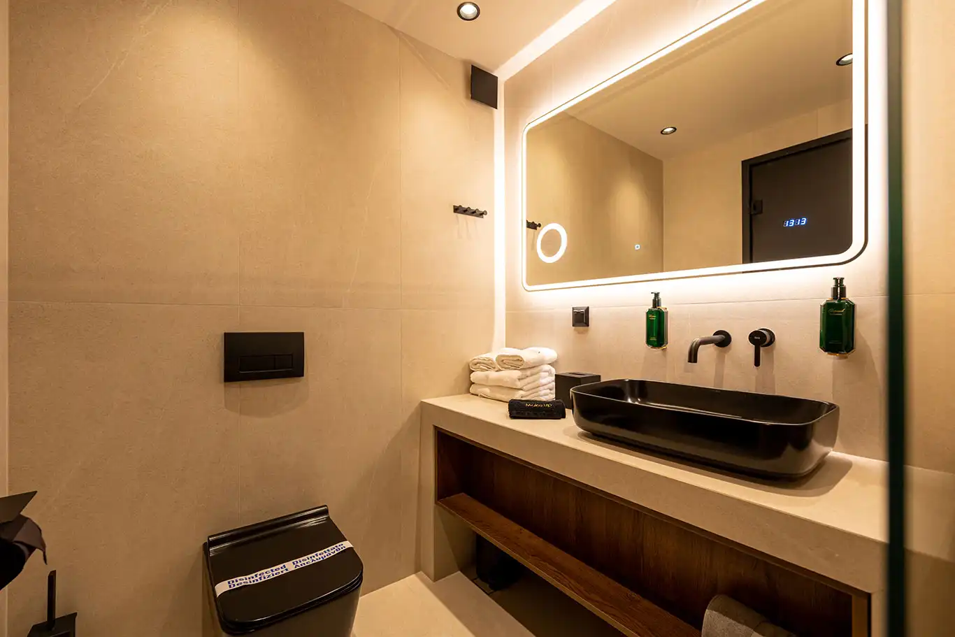 Modern bathroom with black countertop basin, illuminated mirror, folded towels and black wall-mounted toilet