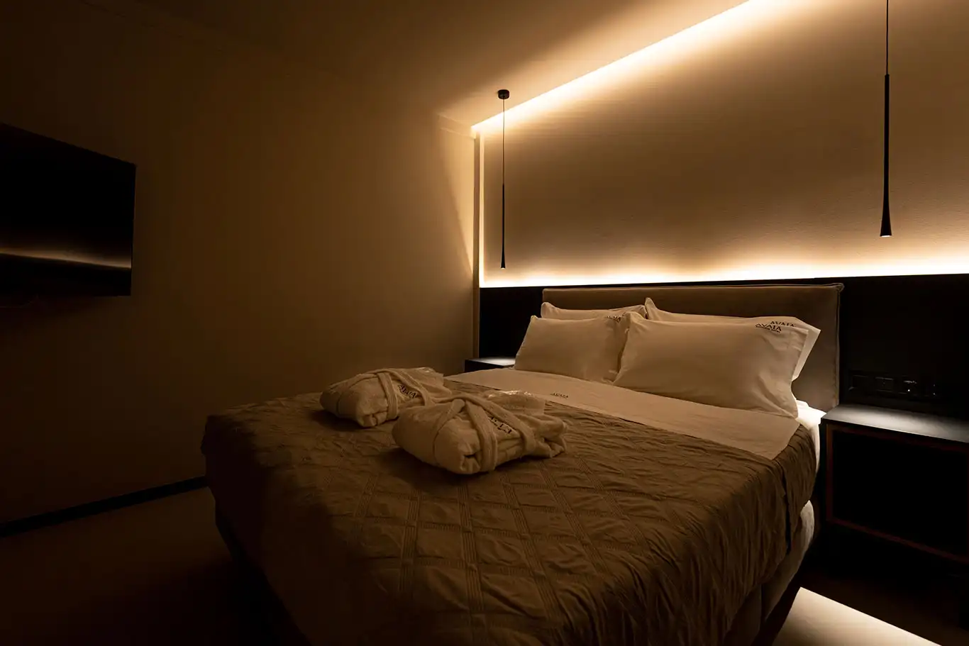 Double bed with white bedding, folded bathrobes, wall-mounted television and illuminated headboard wall