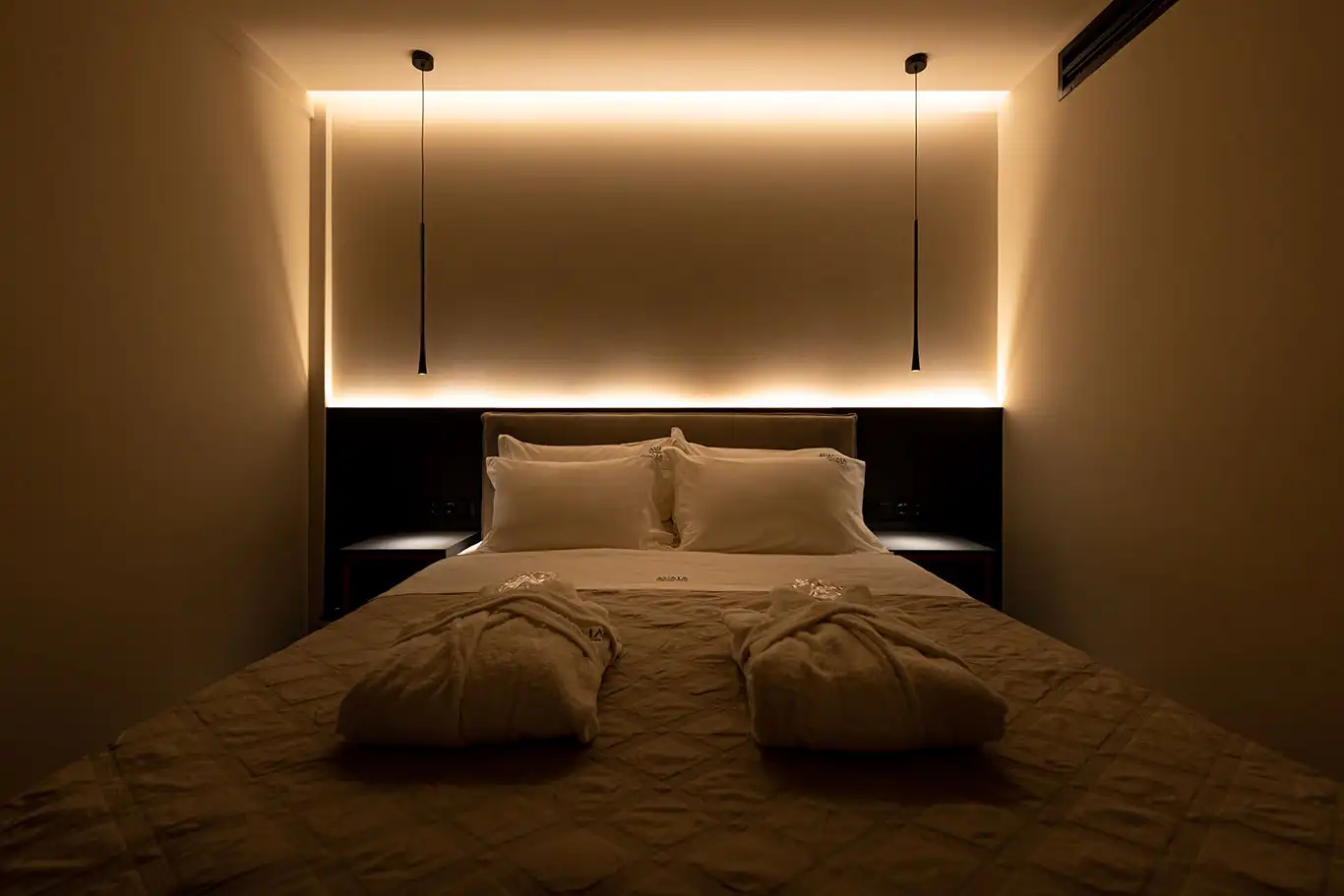 Front view of a double bed with white pillows, folded bathrobes, bedside tables and warm concealed lighting