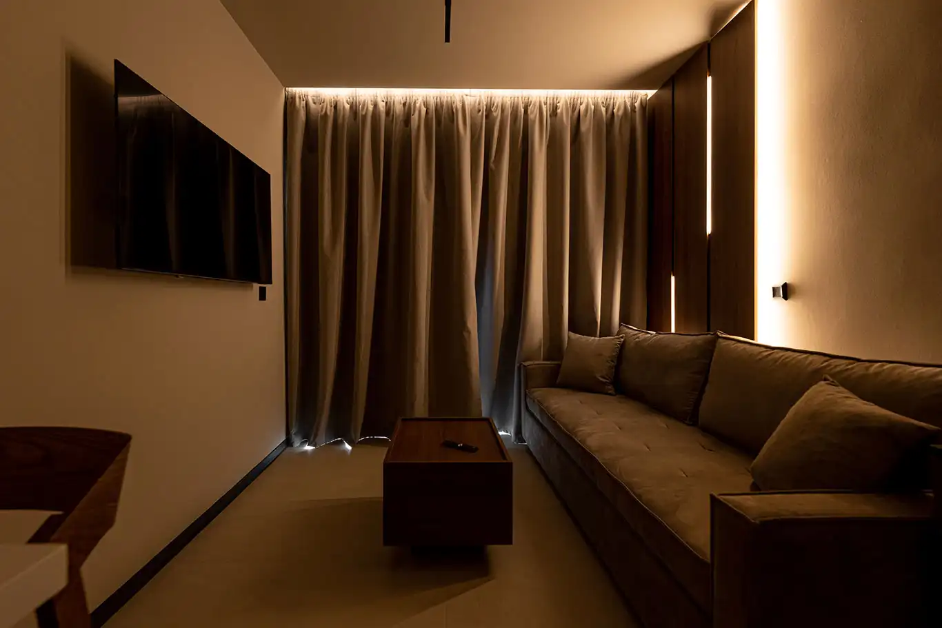 Living area with beige sofa, wooden coffee table, wall-mounted television, dark curtains and illuminated wooden panels
