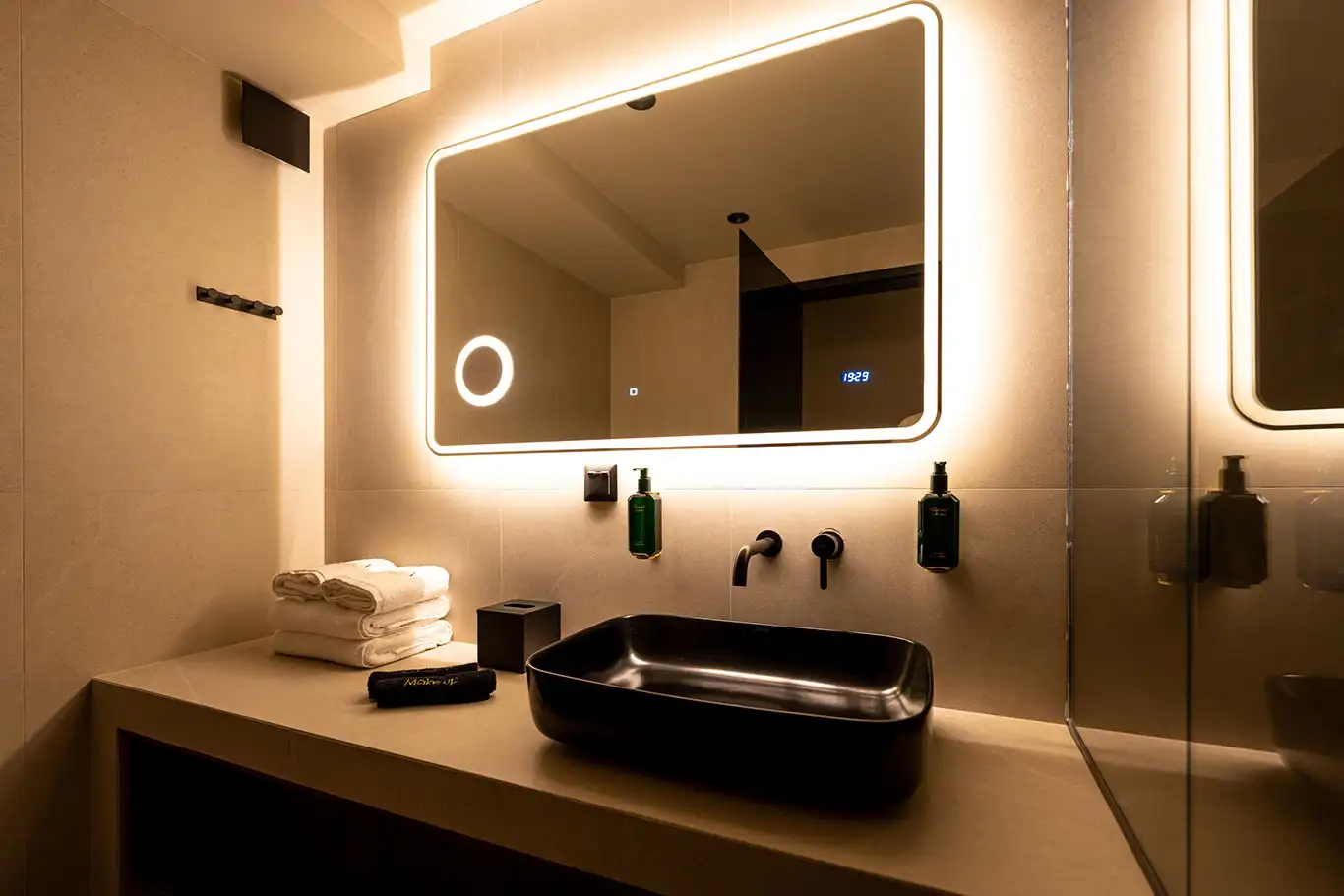 Black countertop basin with wall-mounted tap, illuminated mirror, folded towels and bathroom amenities