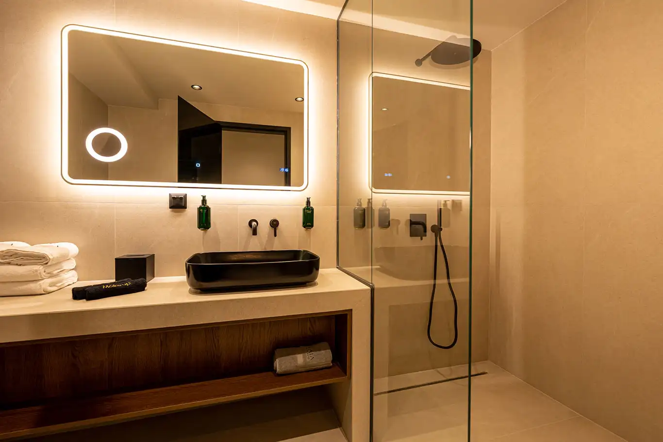Modern bathroom with black basin, illuminated mirror, folded towels and glass walk-in shower with black fixtures