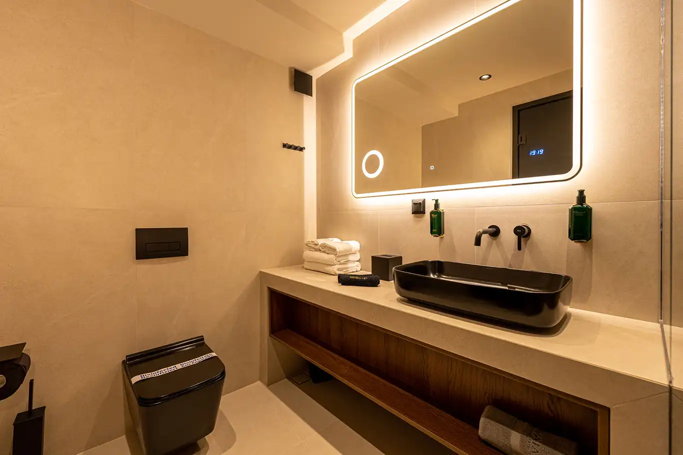Modern bathroom with black countertop basin, illuminated mirror, folded towels and black wall-mounted toilet