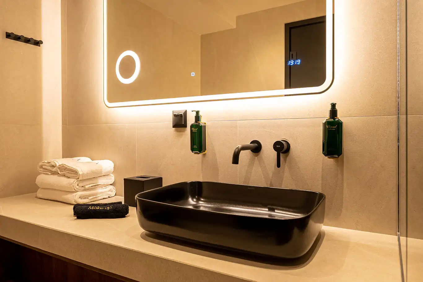 Close-up of a black countertop basin, wall-mounted tap, illuminated mirror, folded towels and toiletries