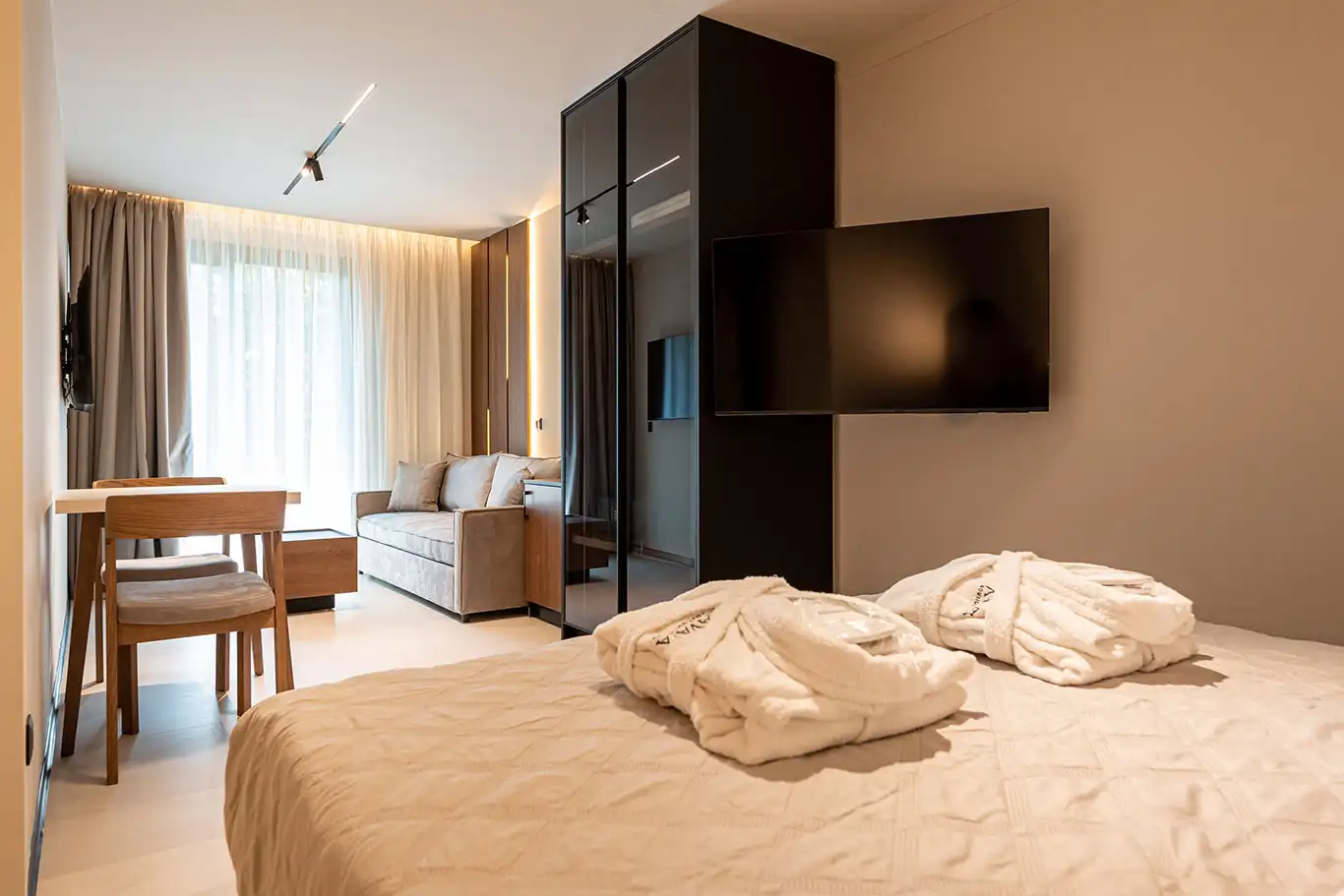 Double bed with folded bathrobes overlooking the dining table, sofa, television and dark glass wardrobe