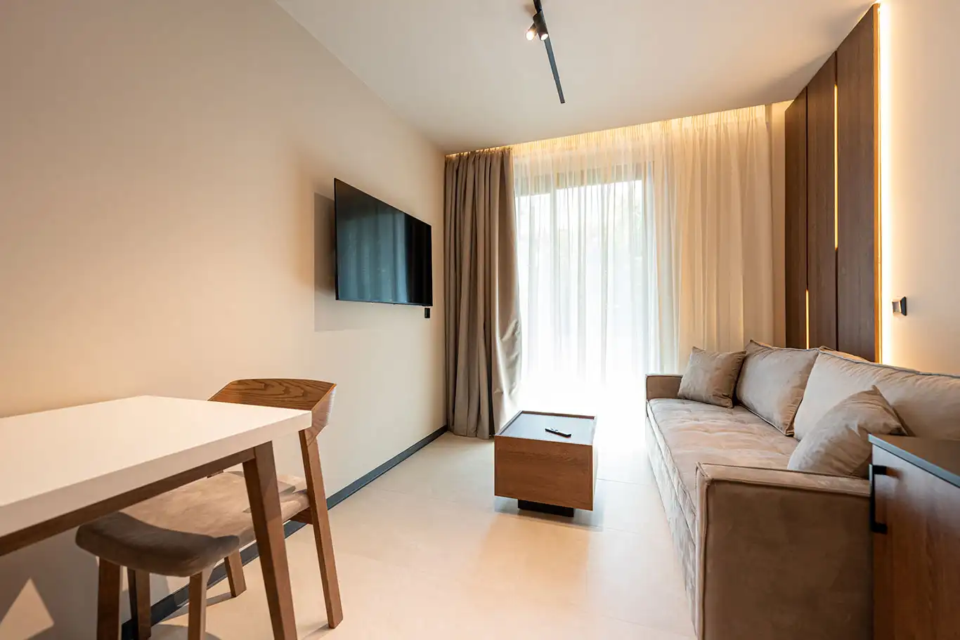 Modern living area with beige sofa, wooden coffee table, white dining table, chairs and wall-mounted television