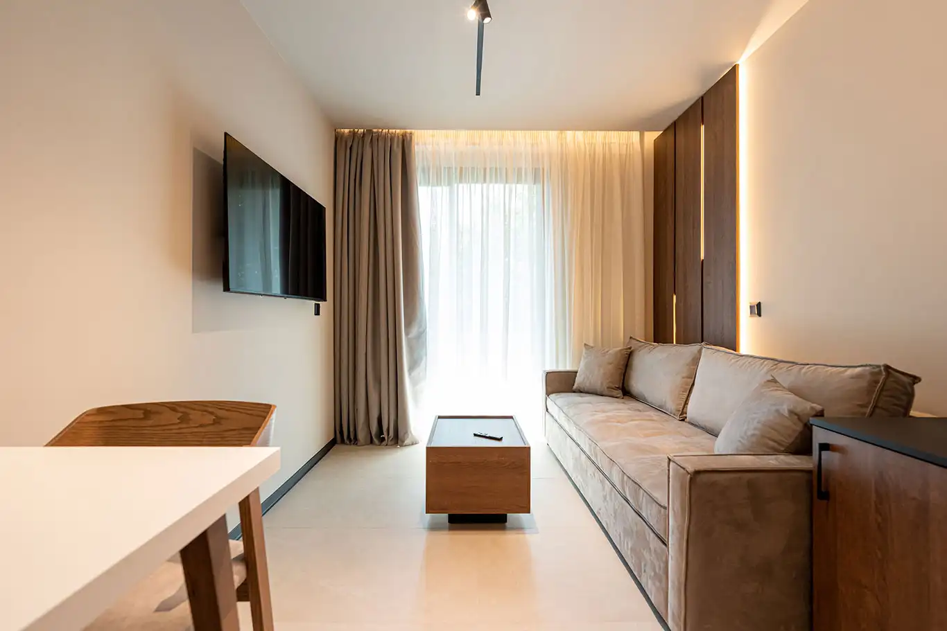 Bright living area with beige sofa, wooden coffee table, wall-mounted television and floor-to-ceiling curtains