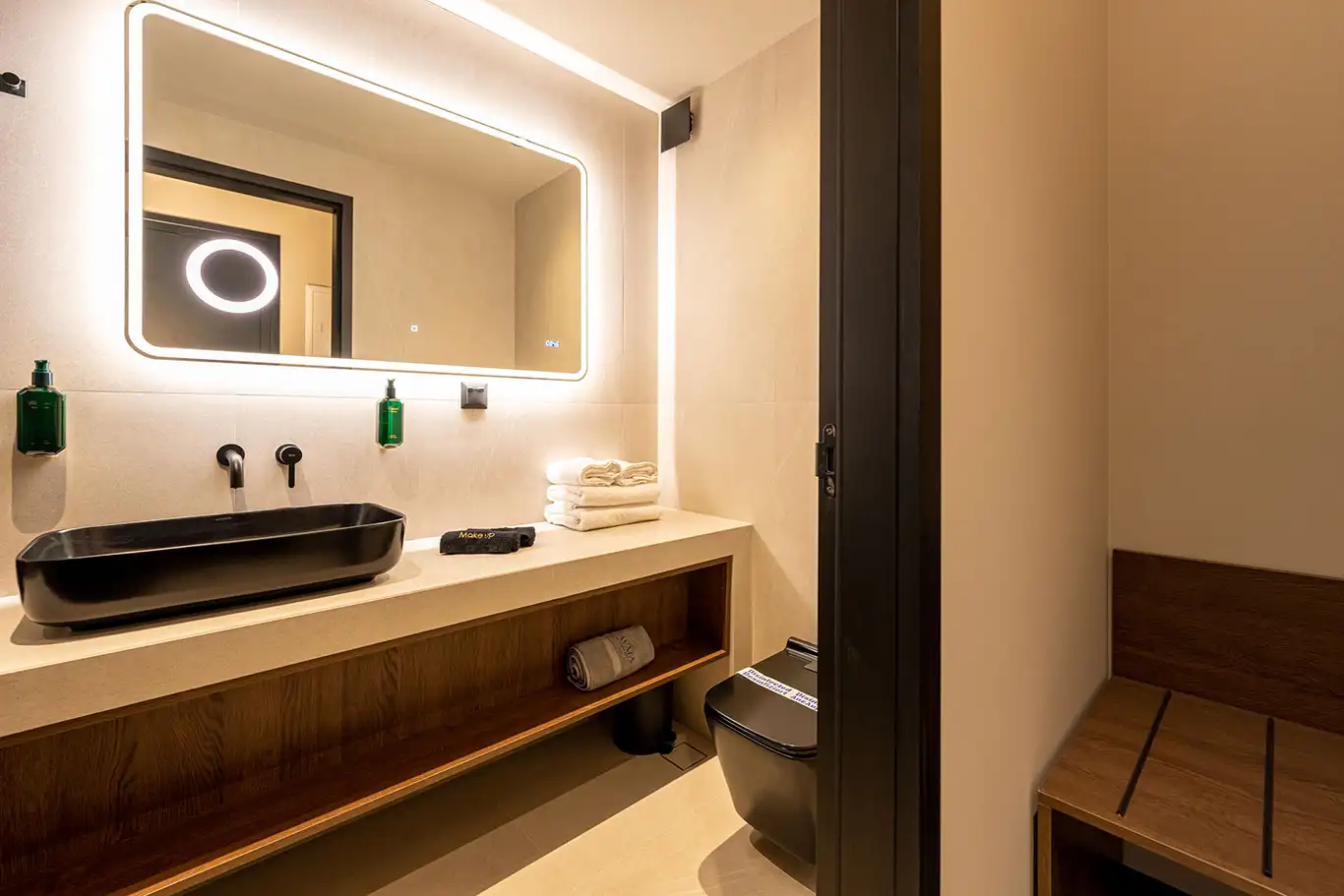 Wide bathroom vanity with black basin, illuminated mirror, towels and adjacent wooden bench