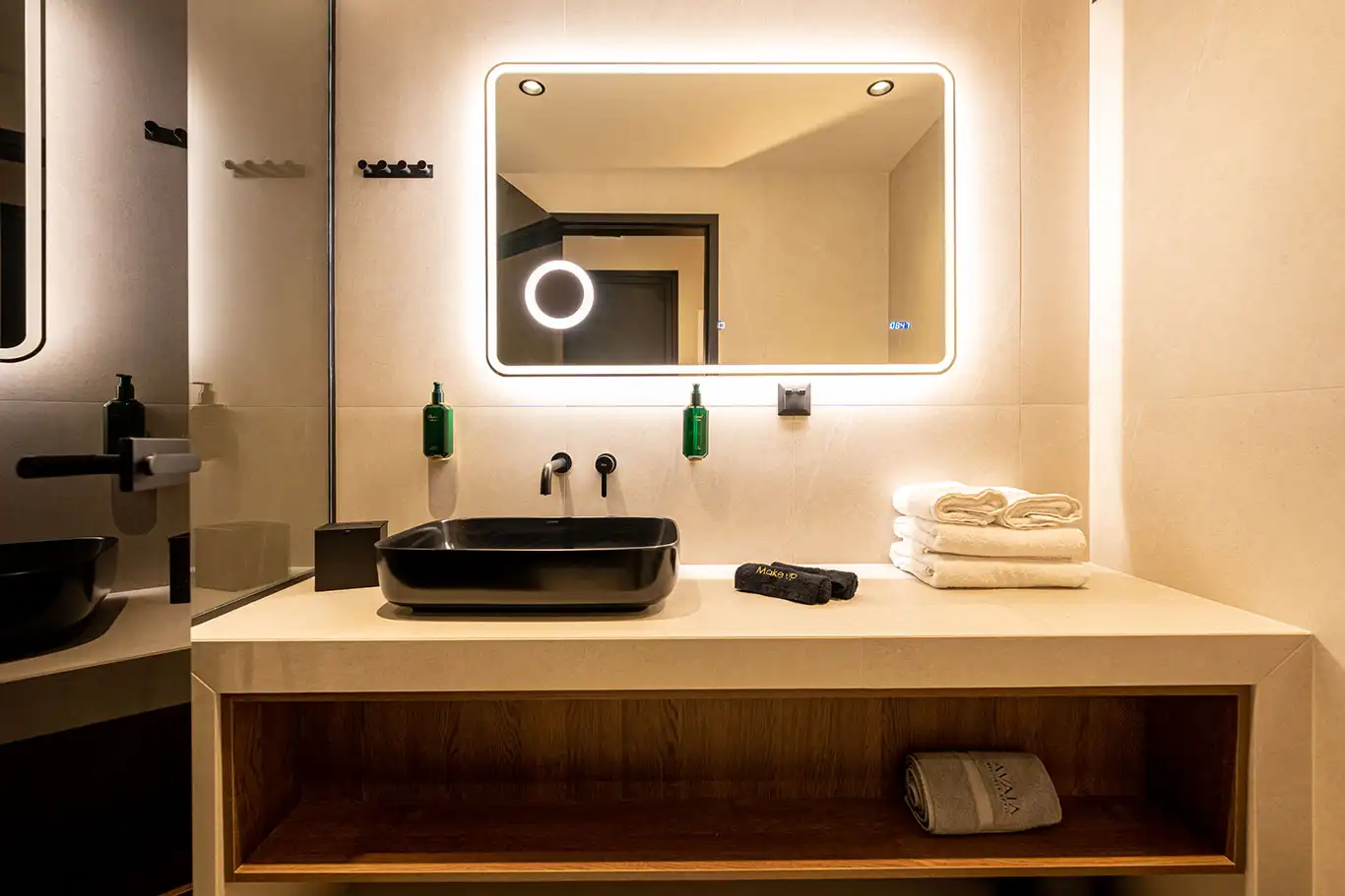 Bathroom vanity with black rectangular basin, illuminated mirror, toiletries and folded towels