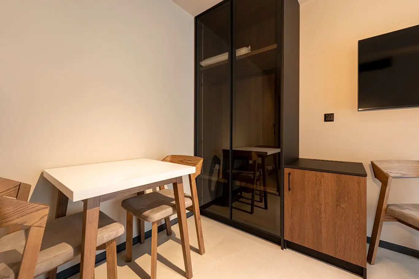 Compact dining table with wooden chairs beside a dark glass wardrobe and storage cabinet