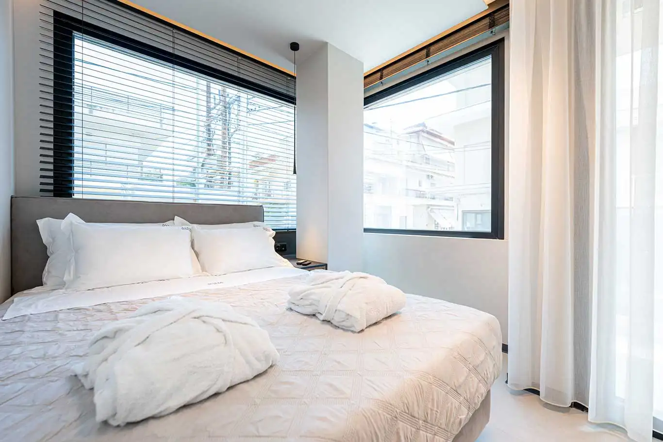 Double bed with white bedding beside large corner windows and sheer curtains