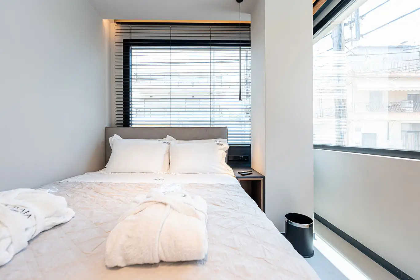 Bright double bedroom with white bedding, bathrobes and large corner windows with blinds