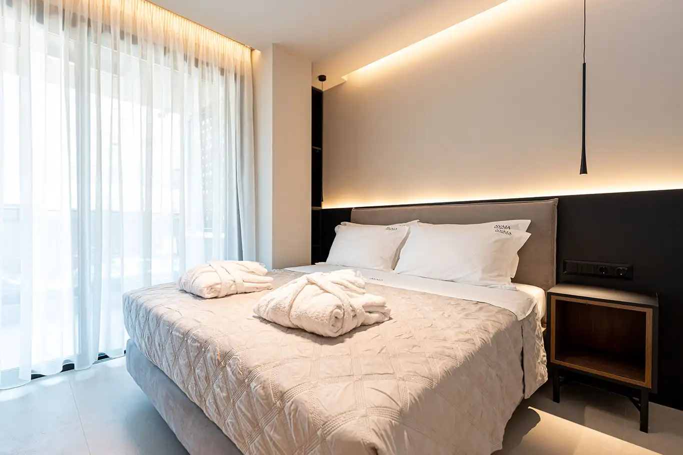 Double bed with white bedding, folded bathrobes, illuminated headboard and floor-to-ceiling sheer curtains