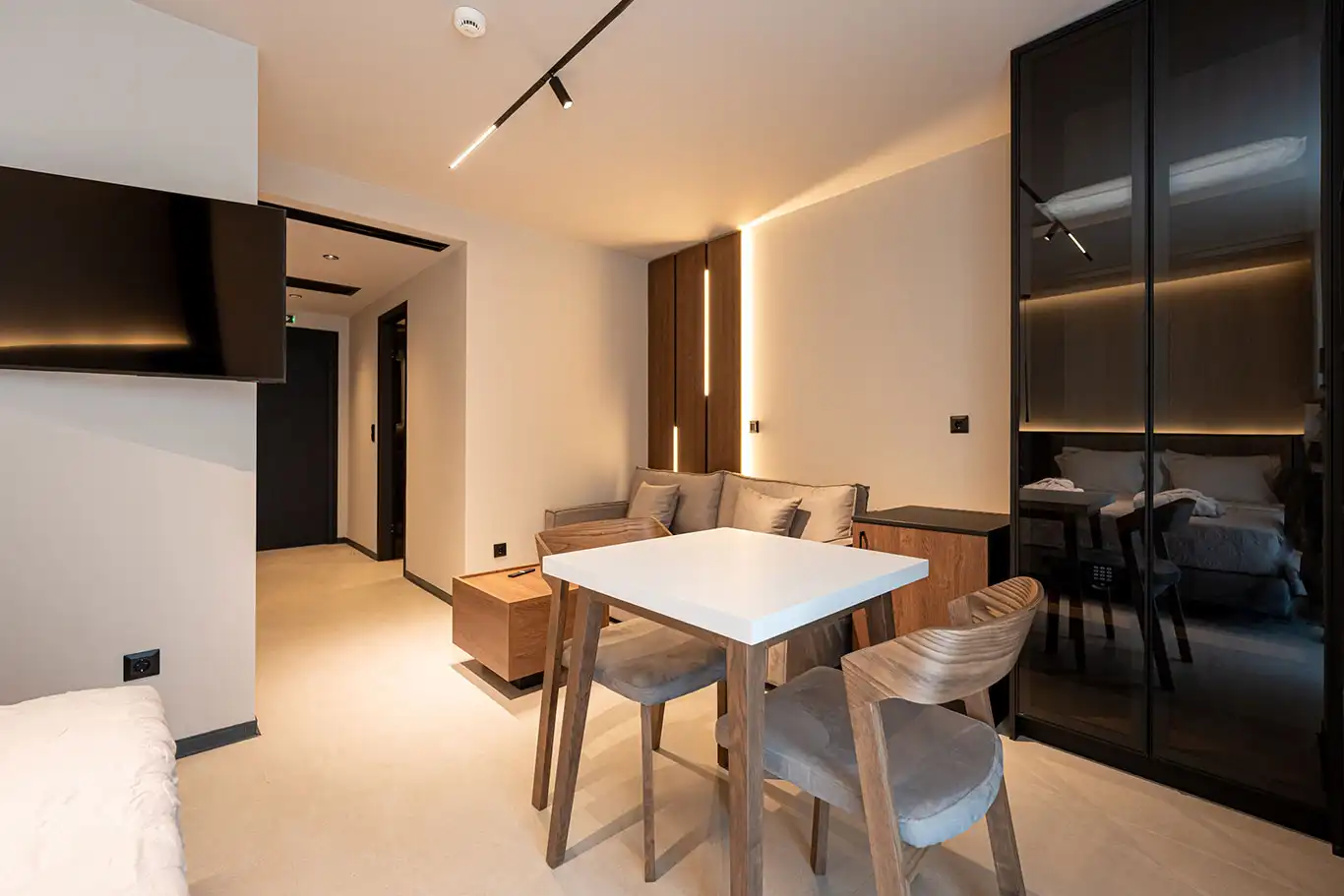 Open-plan room with white dining table, wooden chairs, beige sofa, television and dark glass wardrobe